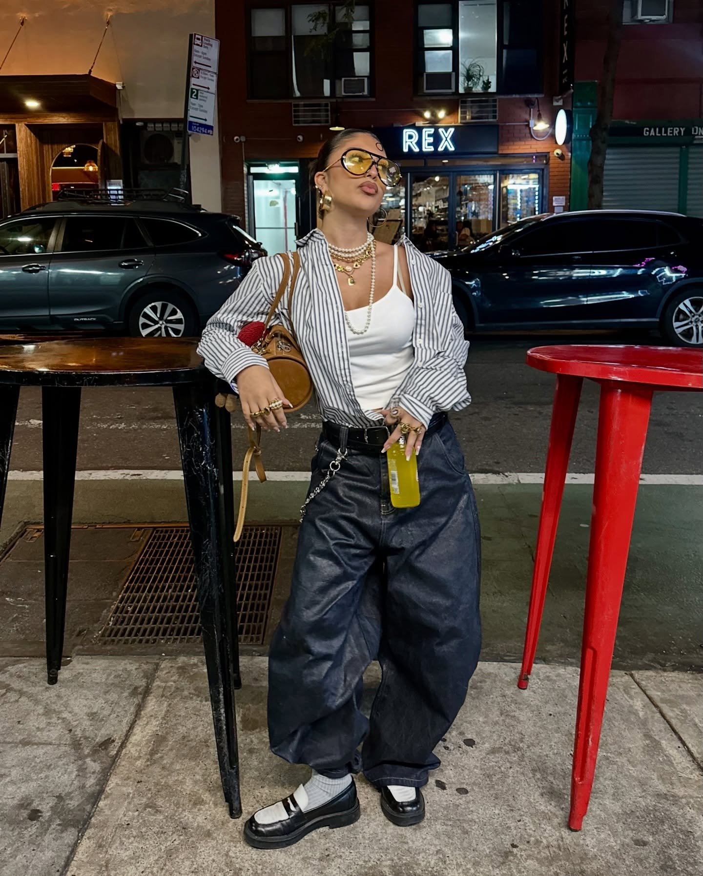 Look com calça jeans baggy escura e regata branca justa, combinados a camisa listrada oversized e acessórios dourados, em