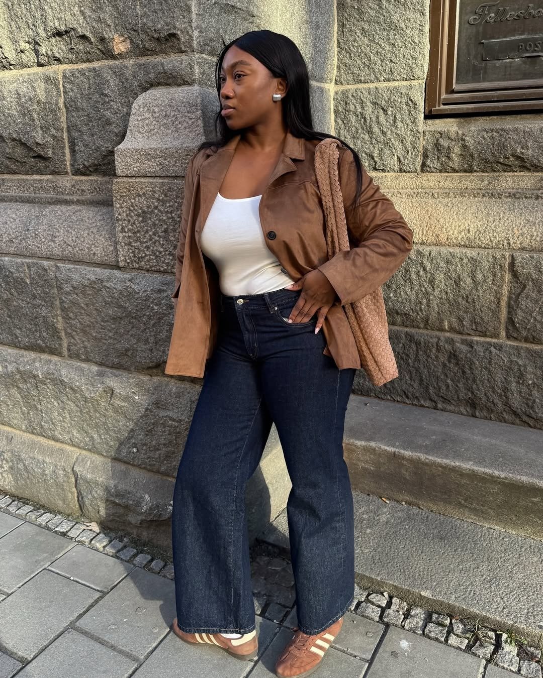 Look com calça jeans flare escura e regata branca justa, combinados com jaqueta marrom de couro e bolsa trançada, estilo