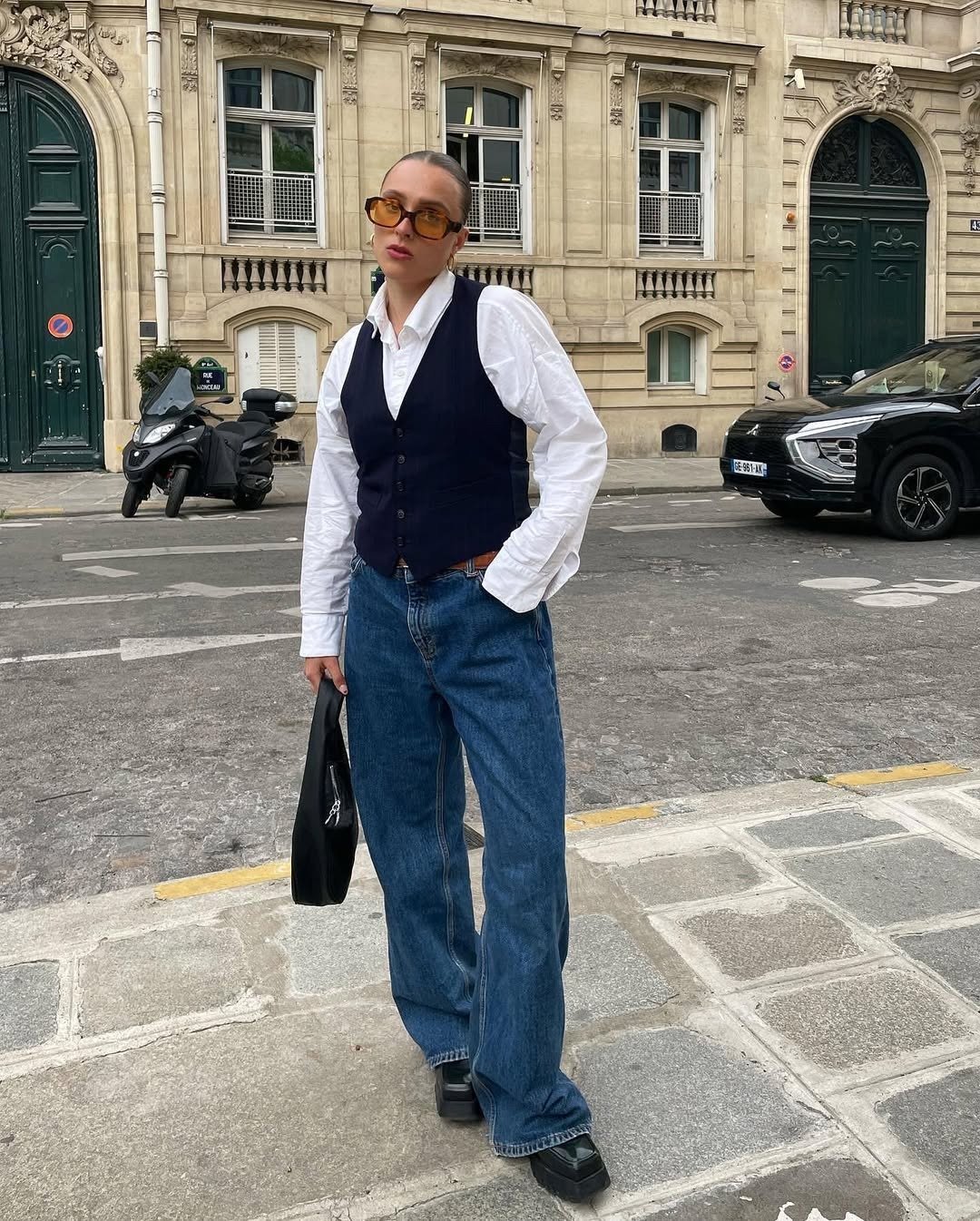 Look com colete preto de modelagem ajustada sobre camisa branca clássica, jeans azul largo, bolsa preta e óculos amarelados,