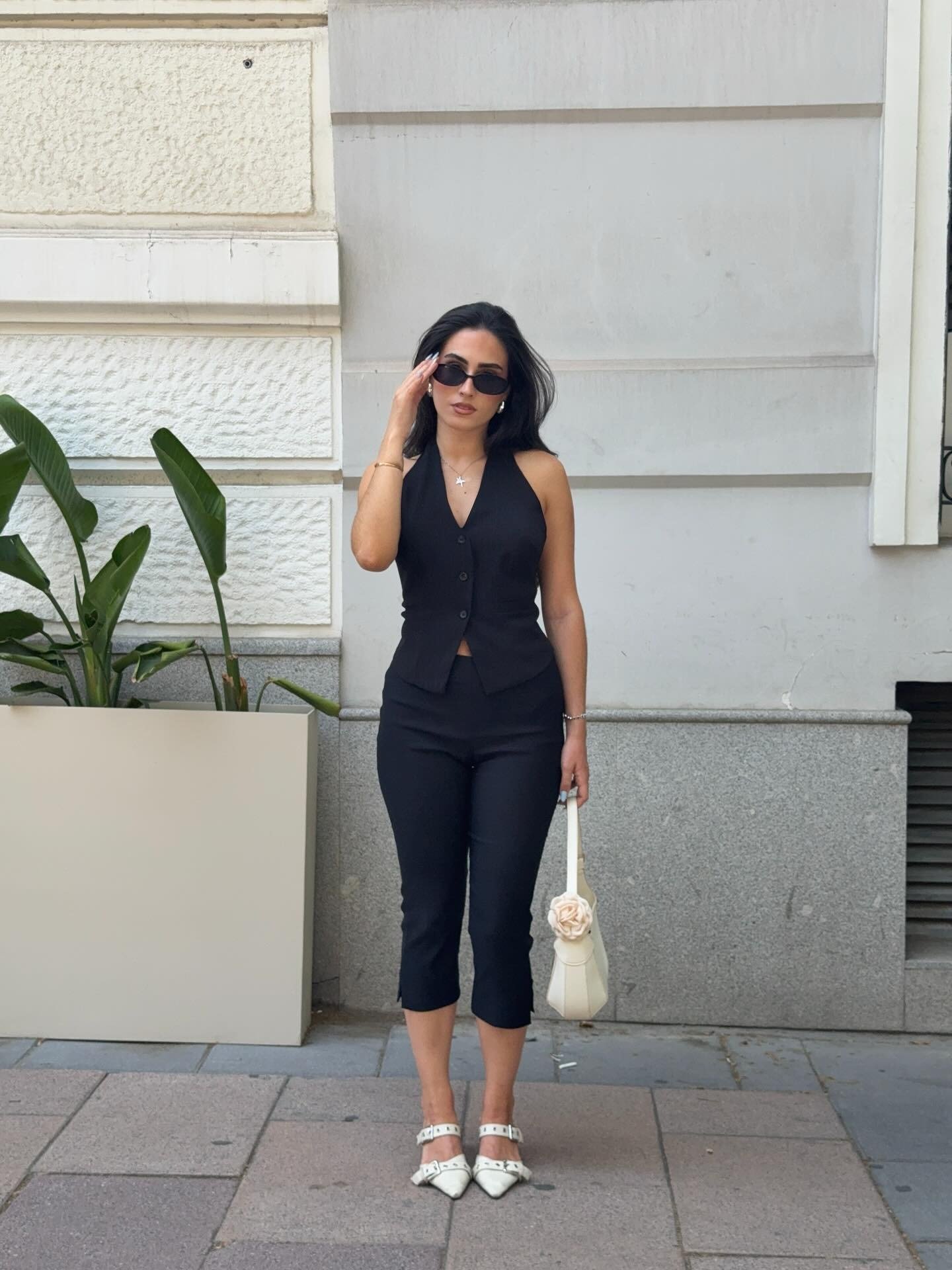 Look com colete preto ajustado de botões, calça cropped justa, acessórios brancos e postura confiante com óculos escuros.