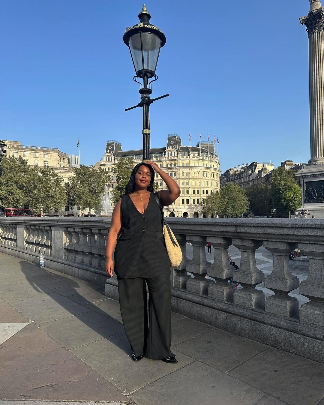 Modelo posa confiante em look com colete preto alongado de alfaiataria risca de giz, calça ampla combinando e bolsa de