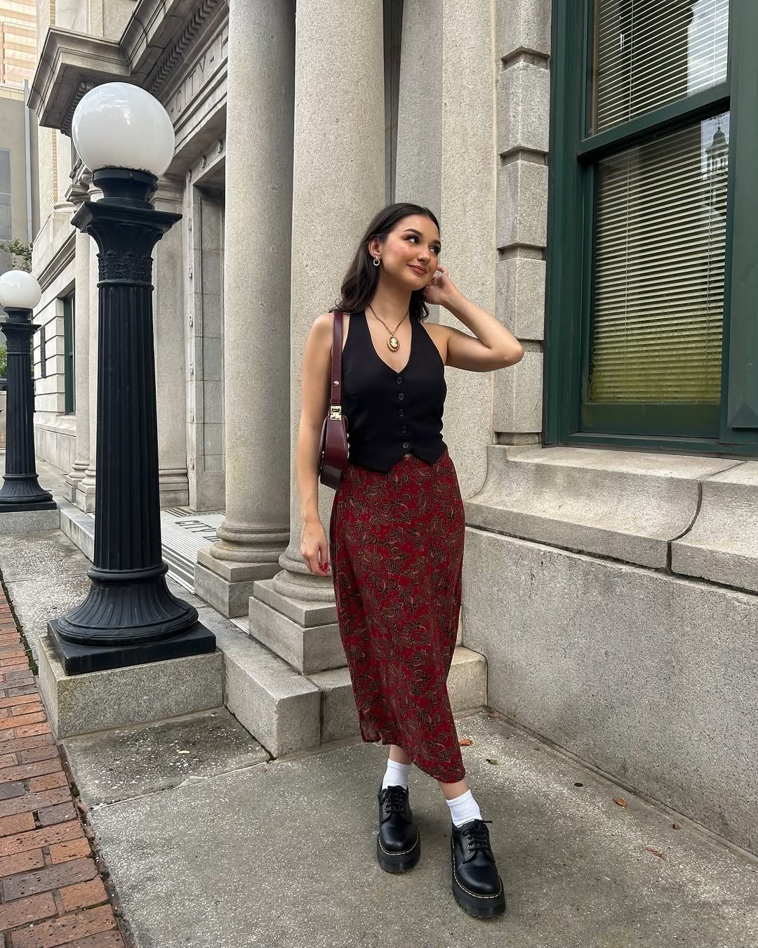 Modelo posa com colete preto ajustado de botões, saia midi vermelha com estampa floral, sapatos pretos robustos e meia