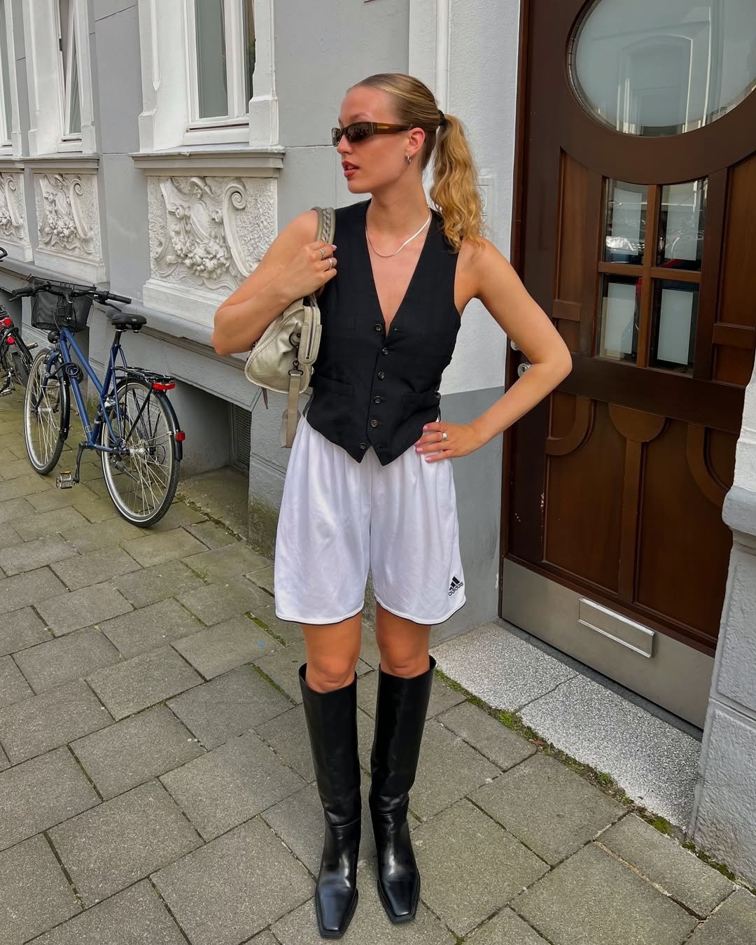 Modelo posa com looks com colete preto de alfaiataria ajustado, short branco esportivo e botas longas de couro, em contraste