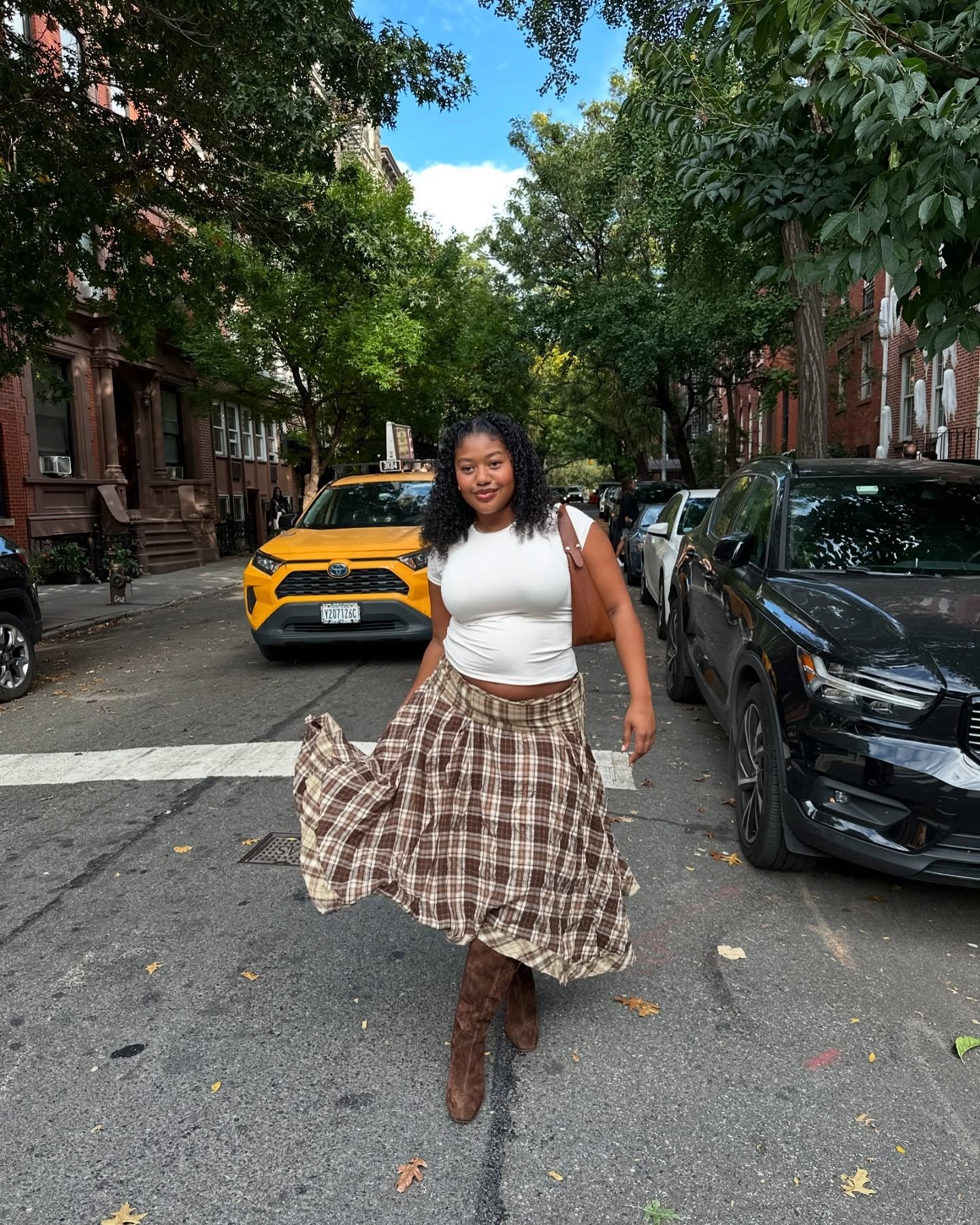 Modelo caminha confiante em rua urbana com saia midi xadrez marrom fluida, camiseta branca justa, bota de camurça e bolsa