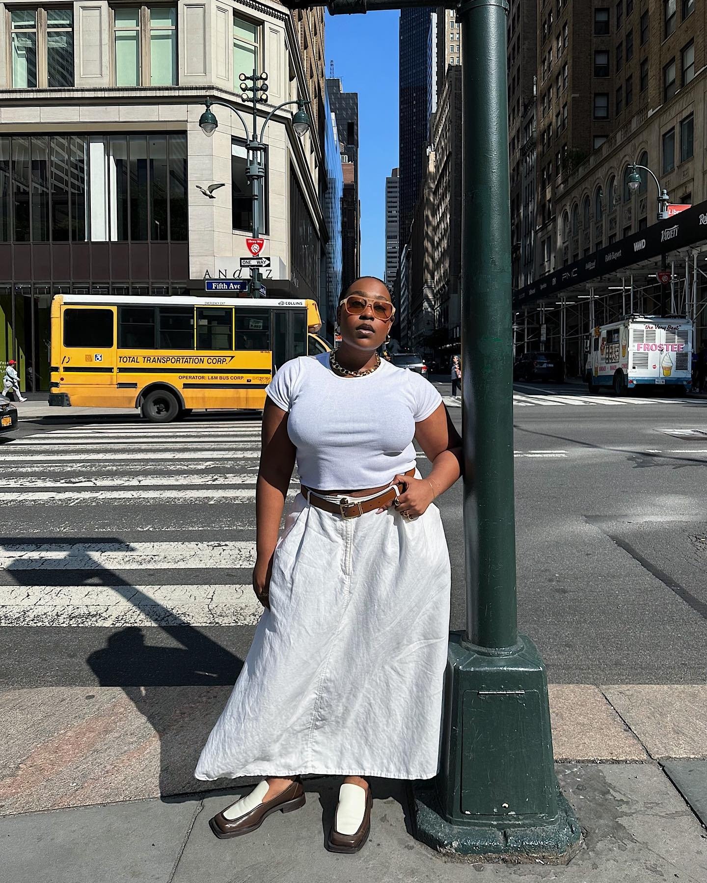 Modelo posa confiante encostada em poste na rua, vestindo cropped branco justo, saia longa de linho branca, cinto marrom e
