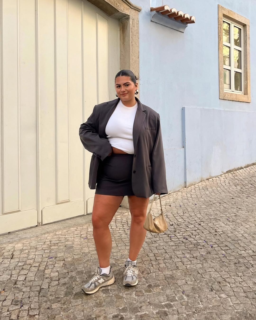 Modelo posa confiante com blazer oversized cinza, saia preta justa, cropped branco e tênis cinza, criando looks fáceis e