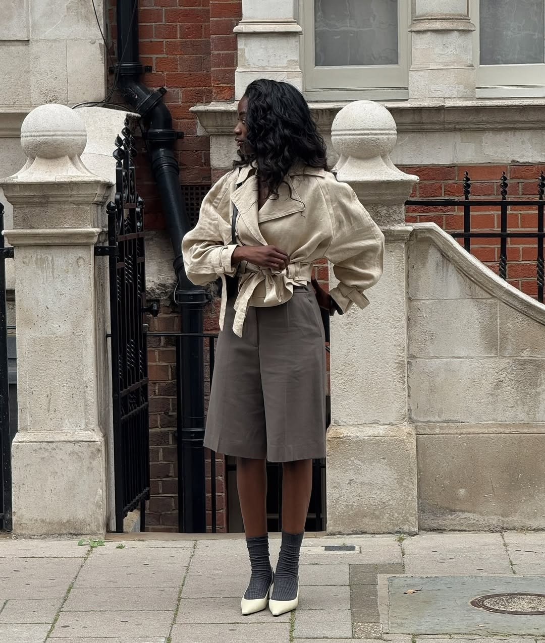 Modelo posa de lado com jaqueta cropped bege de linho amarrada, bermuda ampla cinza e scarpin branco com meia cinza, em