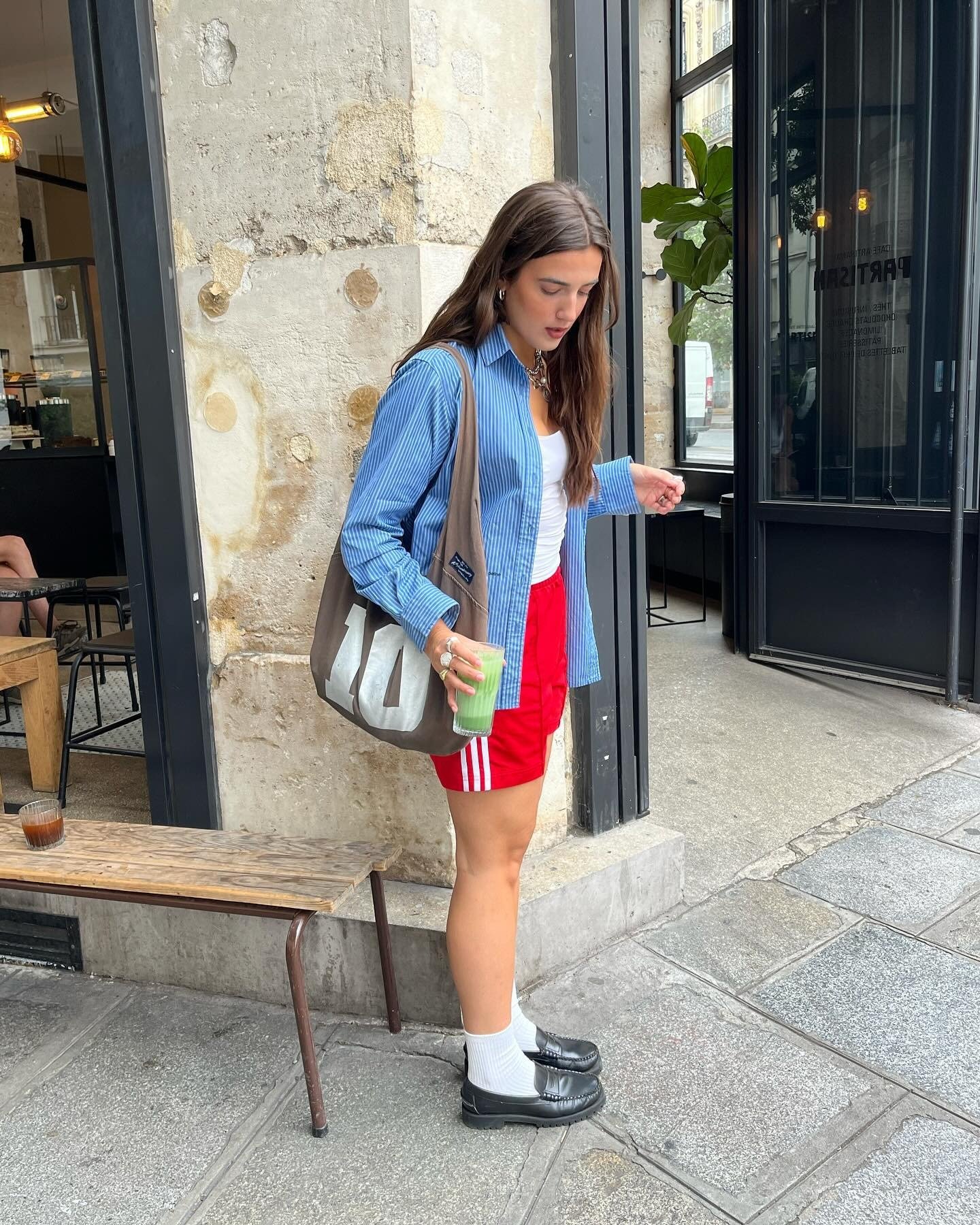 Modelo posa com camisa azul listrada oversized, regata branca, shorts esportivos vermelhos, meias brancas e mocassins