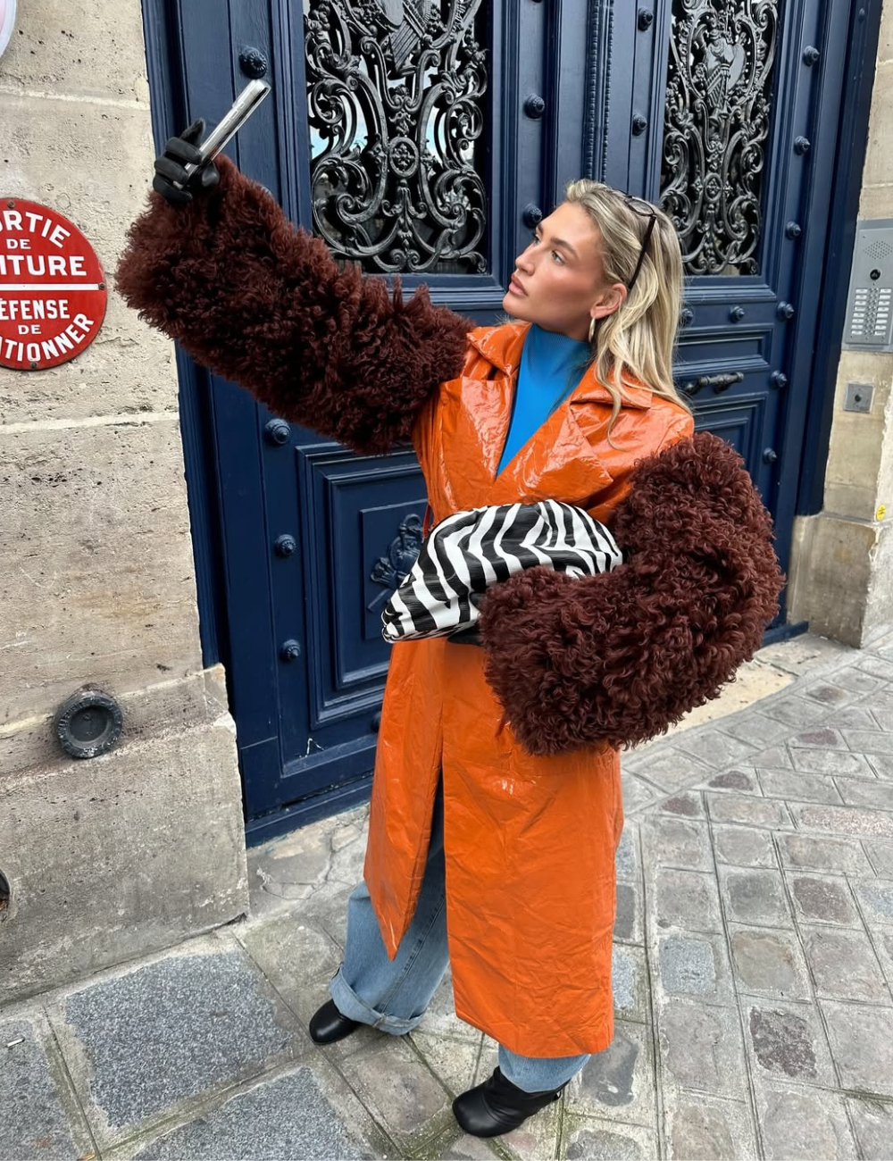 Modelo posa segurando celular e clutch listrada, com casaco laranja vinil e mangas volumosas em pelúcia marrom.