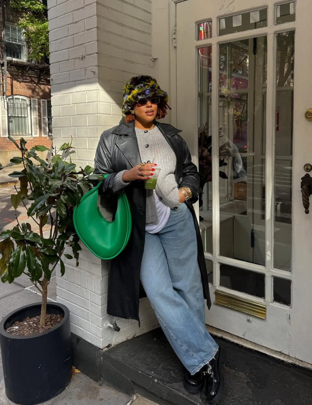 Modelo posa com casaco longo de couro preto, suéter cinza texturizado, jeans largos, bucket hat peludo colorido.
