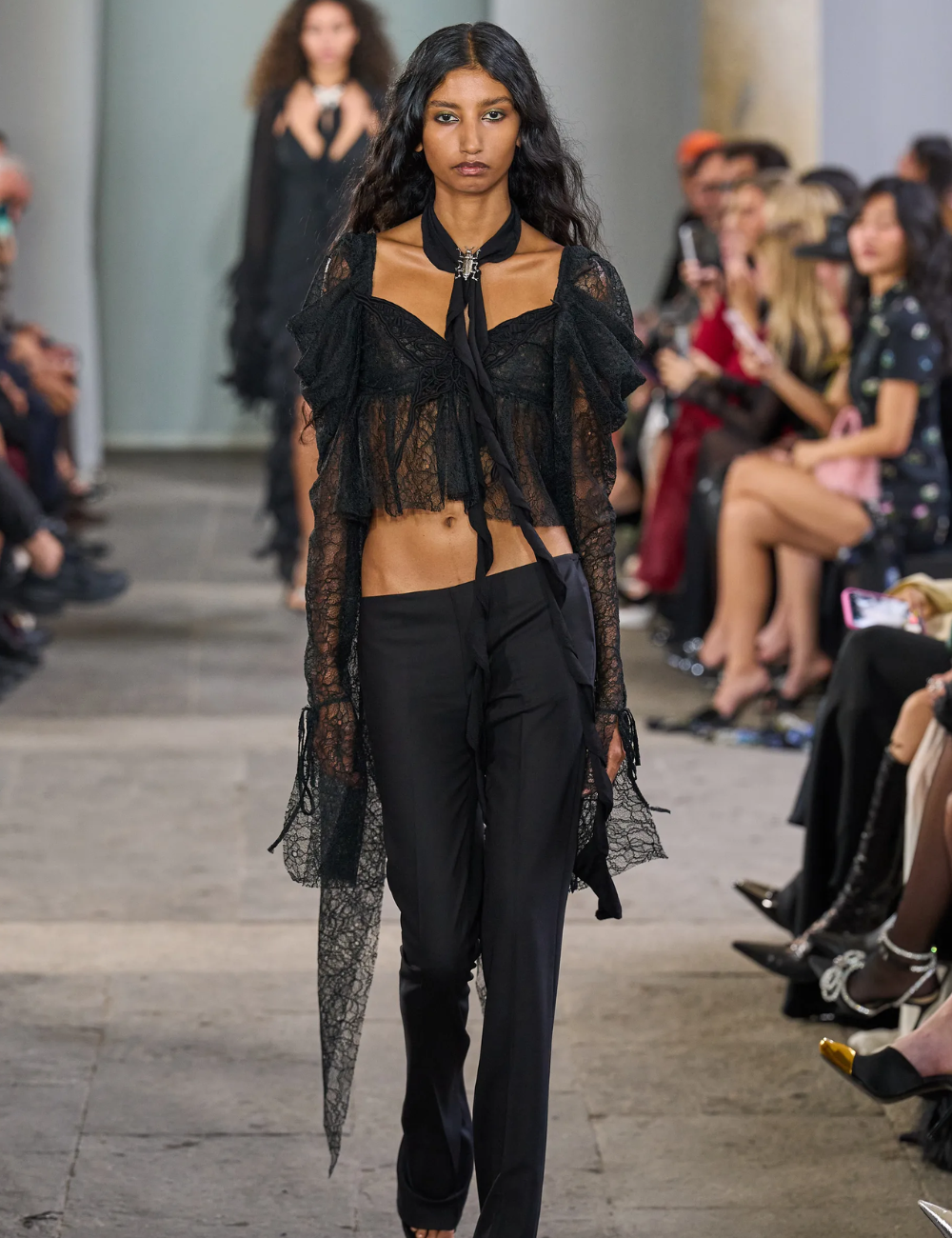 Modelo caminha com postura confiante usando cropped de renda preta com mangas longas e calça reta, tendência de 2026.