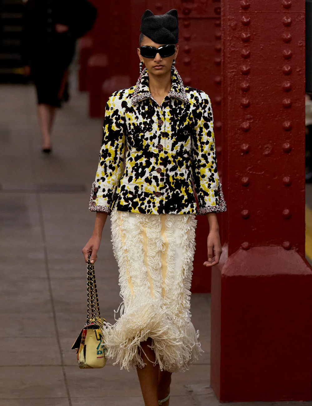 Modelo posa com blazer estampado em preto, amarelo e branco, saia texturizada com franjas e acessórios elegantes.