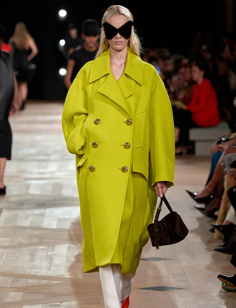 Modelo posa com casaco oversized amarelo-limão em tecido estruturado, combinando calça branca e bolsa marrom.