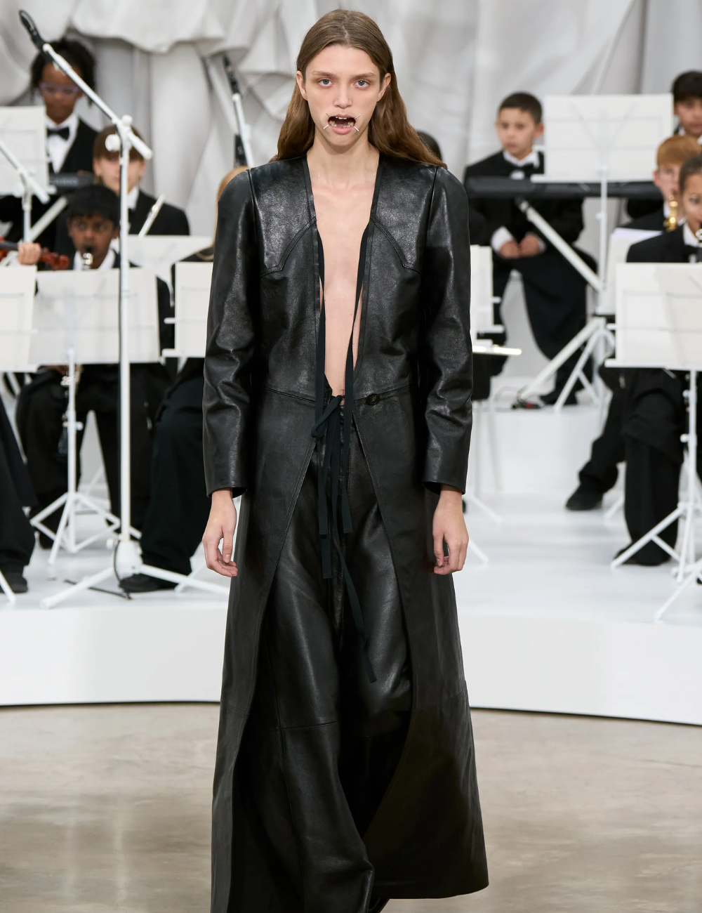 Modelo posa com casaco longo e calça de couro preto, modelagem ampla e minimalista, destacando tendências de 2026.