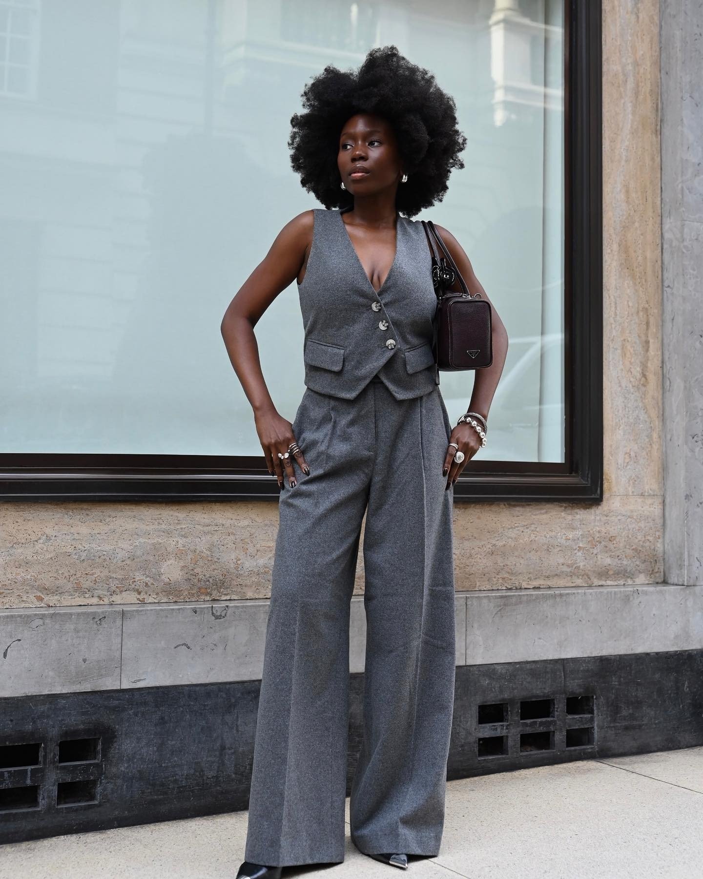 Modelo posa confiante com colete acinturado e calça pantalona cinza de tecido estruturado, combinando bolsa preta e