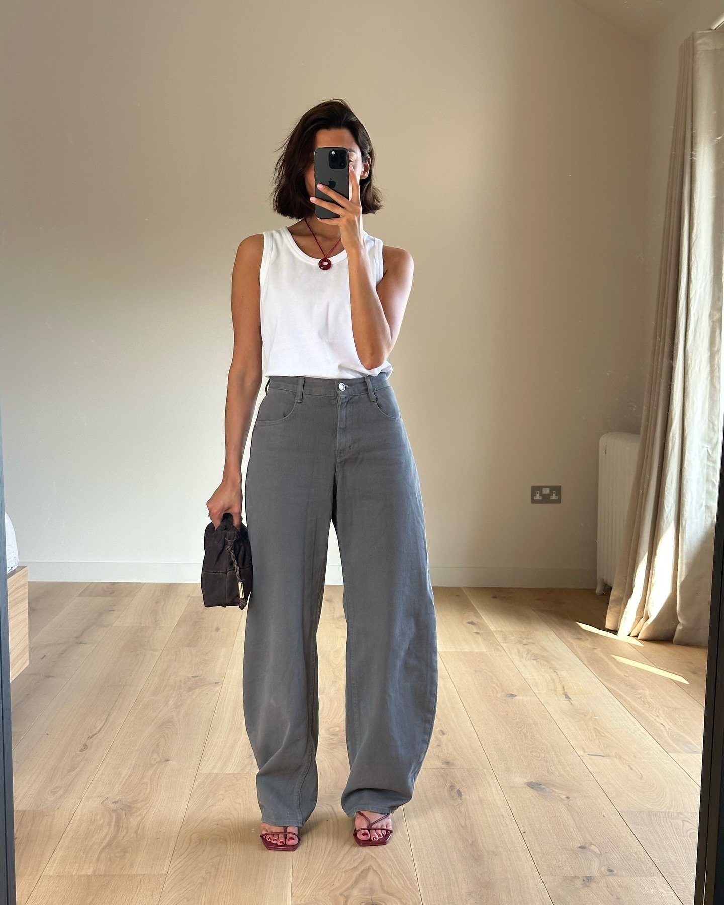 Modelo posa em ambiente neutro com regata branca justa, calça cinza de modelagem ampla e sapato vinho de tiras finas,