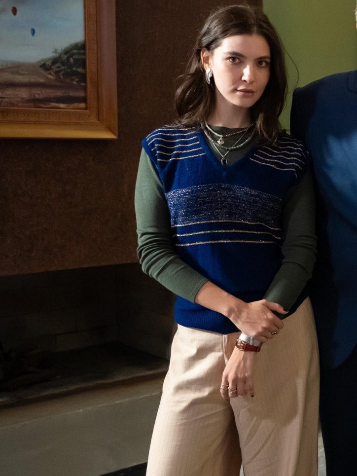 Lorena veste colete azul de tricô com listras douradas sobre blusa verde manga longa e calça bege, posando com expressão