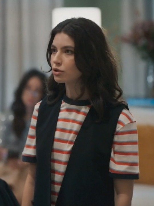 Lorena veste camiseta listrada em branco, vermelho e azul com colete preto, expressando foco e determinação em ambiente