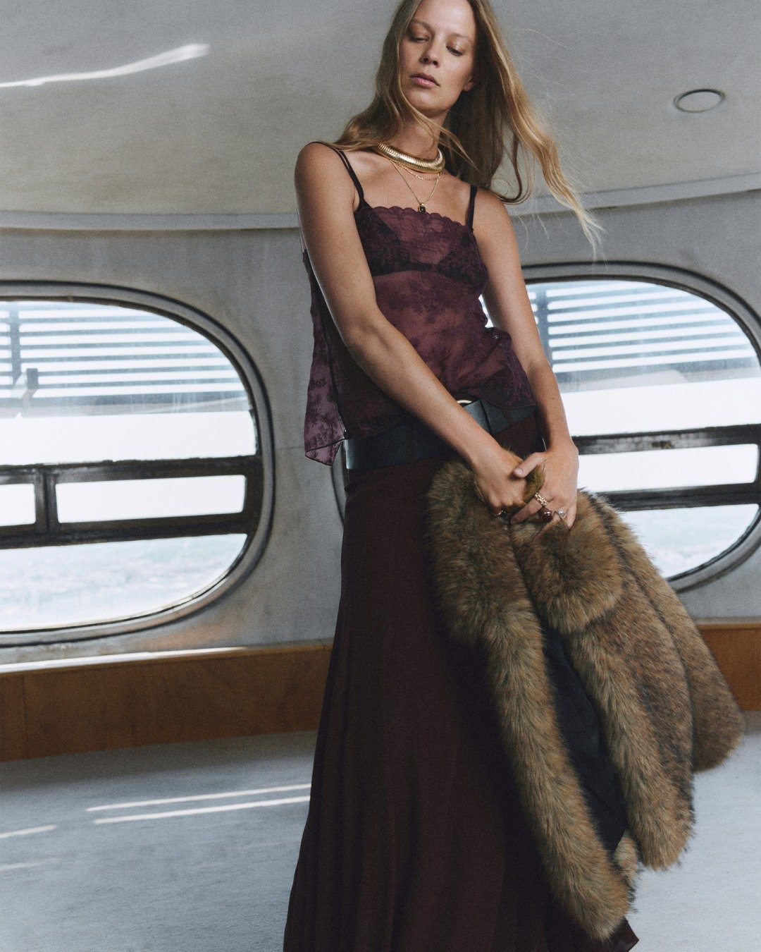 Modelo posa com blusa de renda vinho translúcida, saia longa fluida marrom e casaco de pele marrom, refletindo a indústria