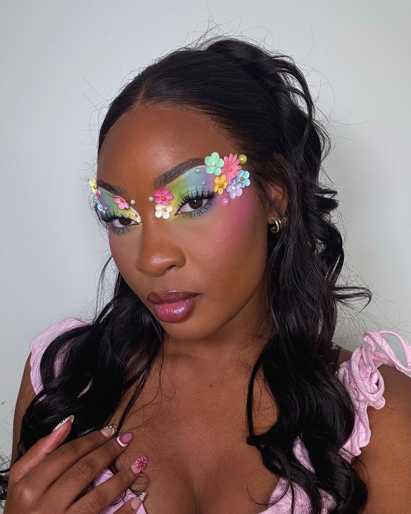 Modelo posa com maquiagem colorida em tons pastel e flores 3D ao redor dos olhos, combinando com blusa rosa de amarração