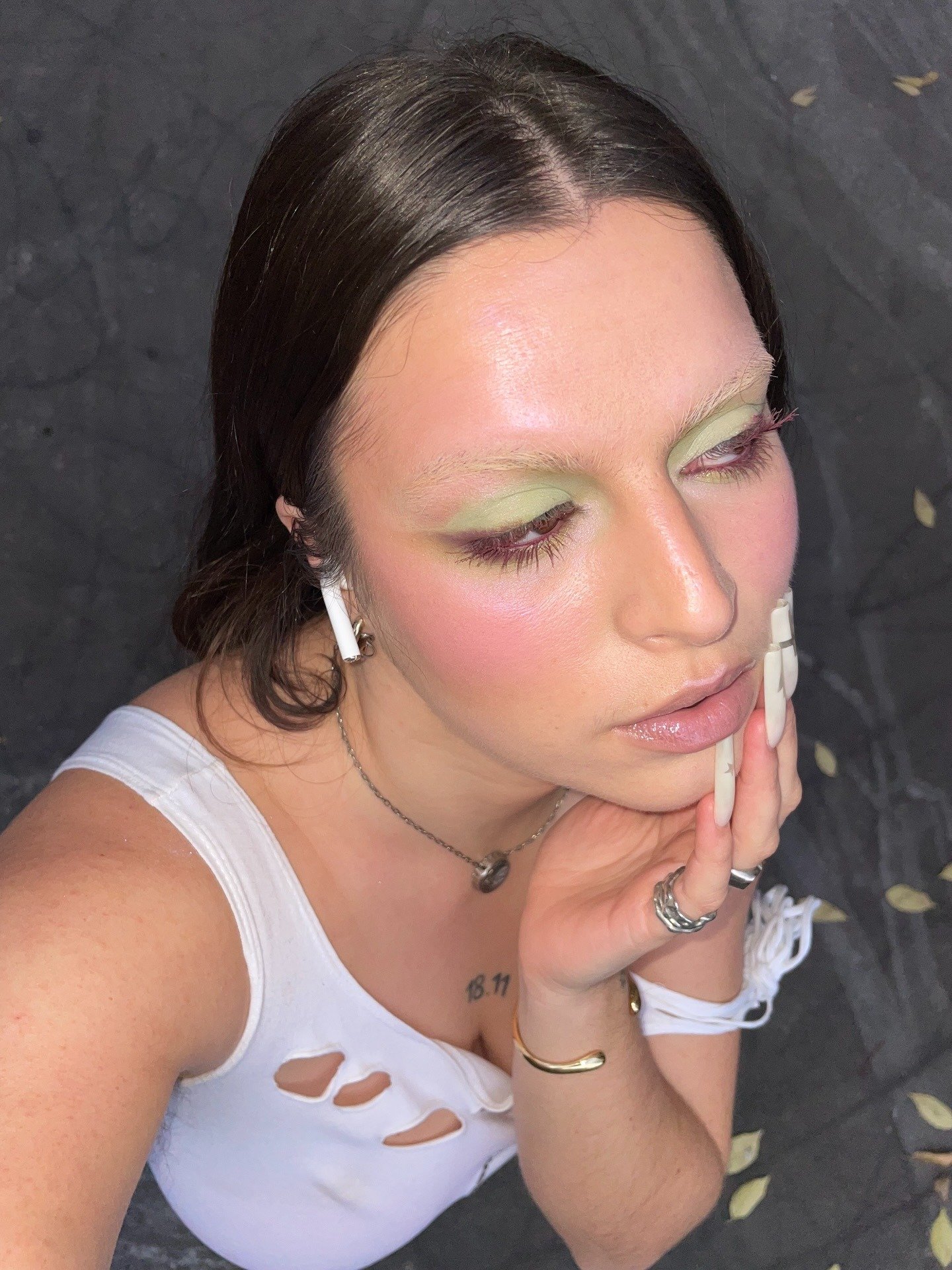 Modelo posa com maquiagem colorida em tons de verde e rosa, unhas longas brancas e blusa branca rasgada, em clima delicado