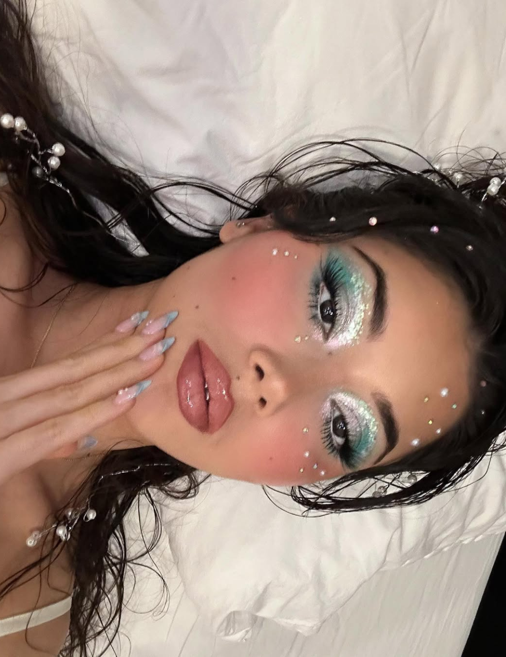 Modelo deitada exibe maquiagens de Carnaval com sombra azul e prata cintilante, blush rosado, gloss nos lábios e cristais.