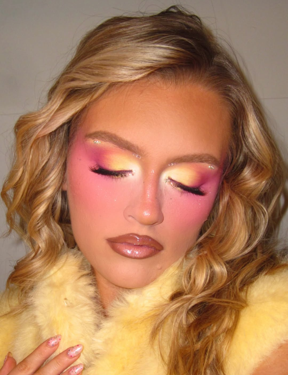 Modelo com olhos fechados exibe maquiagens de Carnaval em tons vibrantes de amarelo, rosa e roxo, com glitter nos olhos.