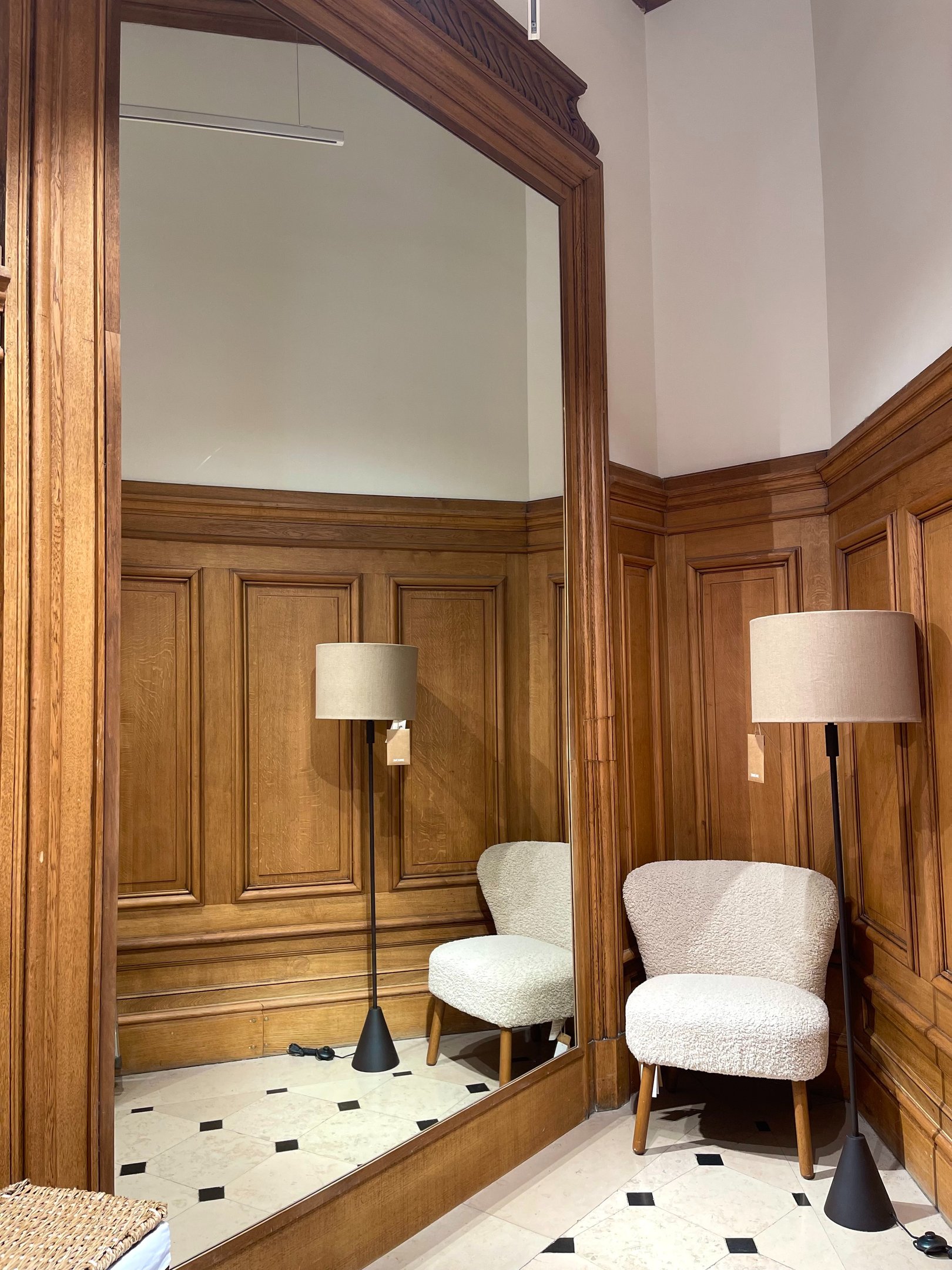 Espelho em casa com moldura de madeira clássica reflete cadeira estofada branca e luminária de piso preta