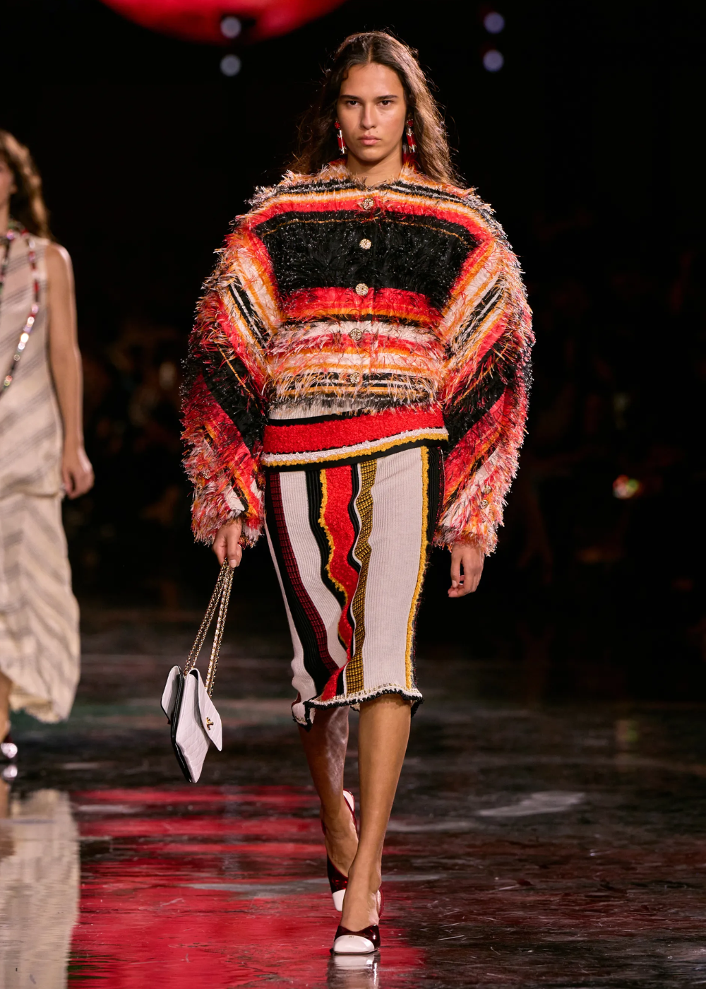 Modelo caminha com postura confiante usando conjunto maximalista de tricô texturizado em listras vermelhas, pretas, amarelas.