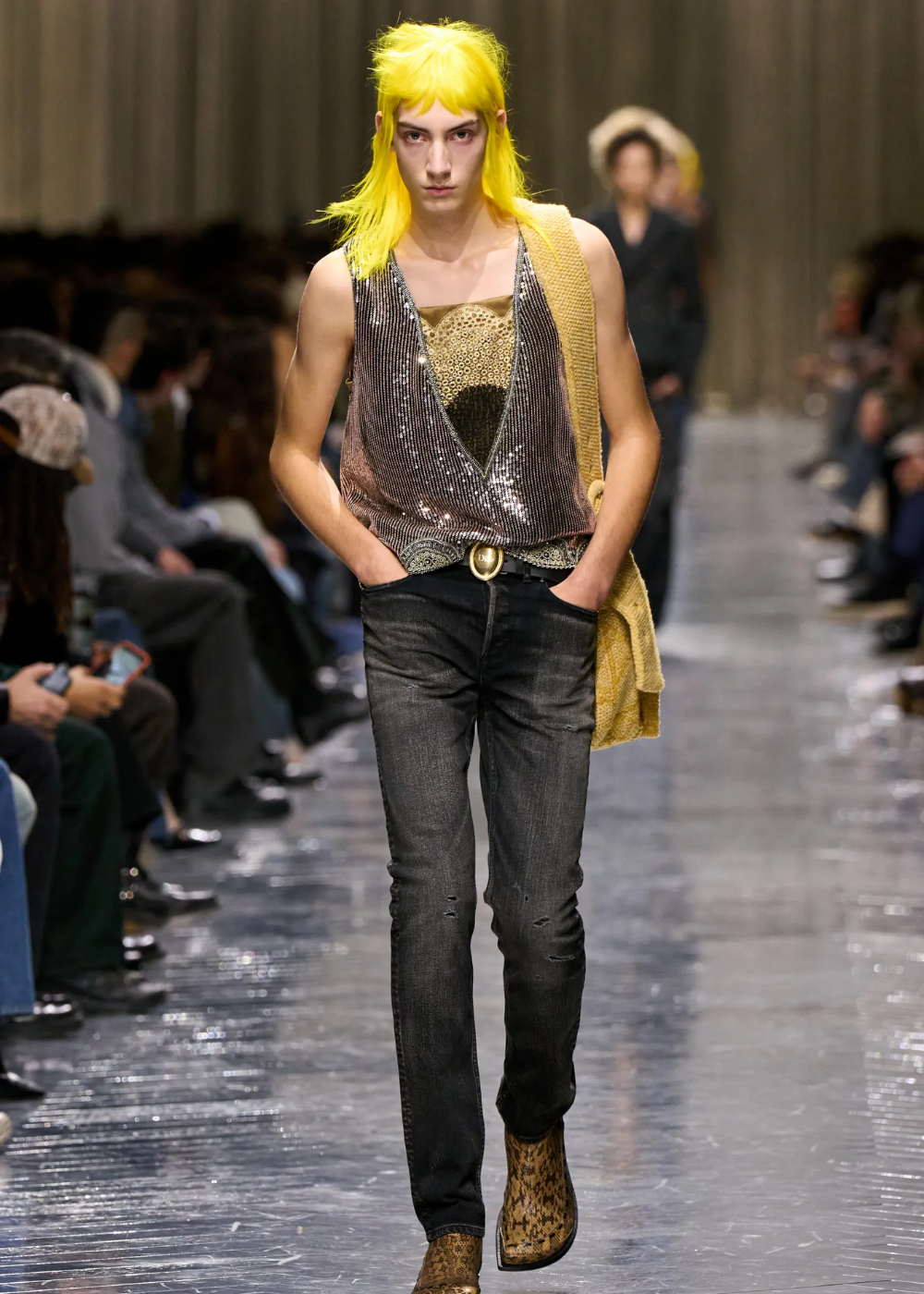 Modelo caminha com postura confiante usando colete prateado brilhante, blusa dourada, jeans escuro e botas animal print.