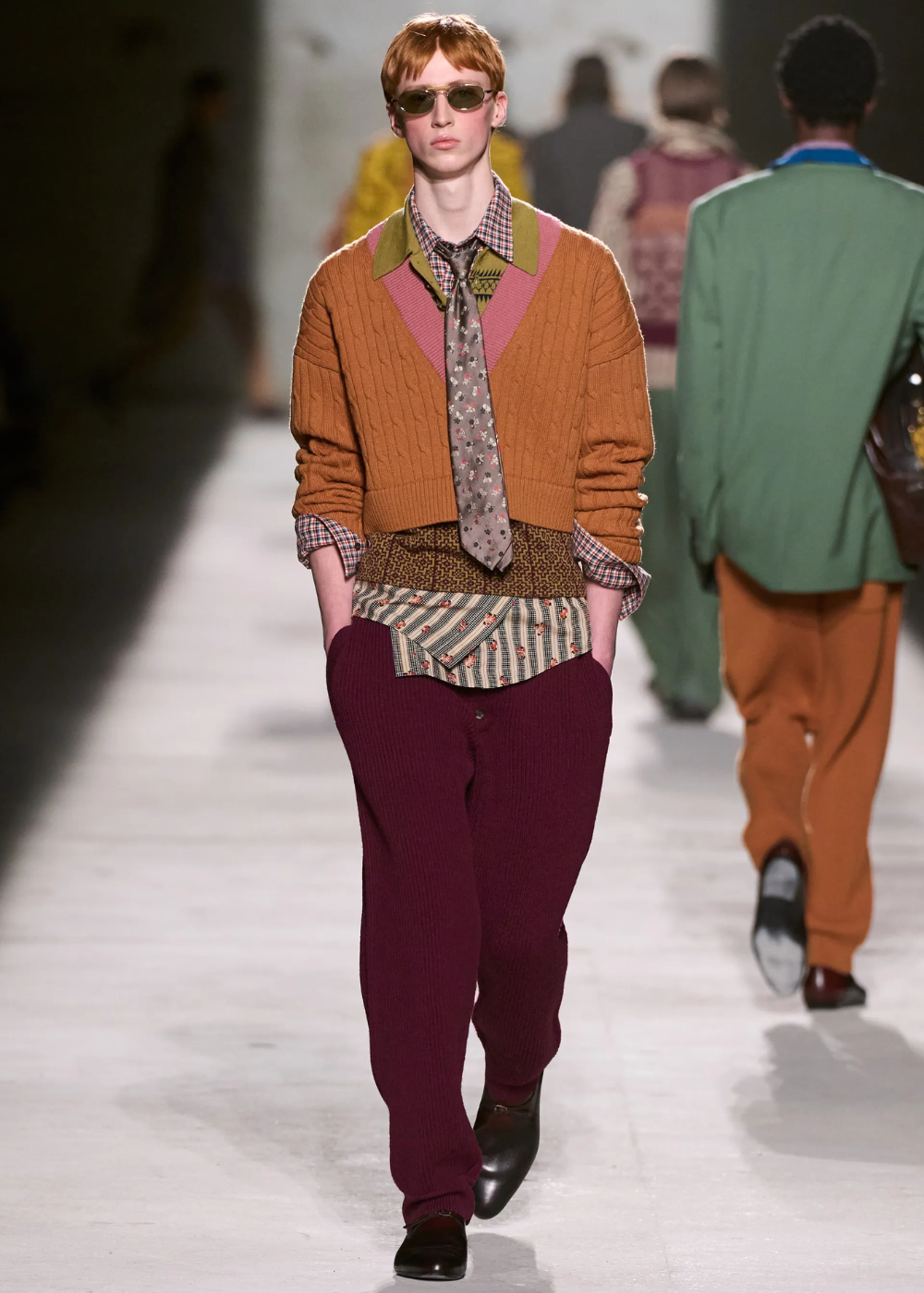 Modelo caminha com postura confiante usando maximalismo em mix de malhas e tecidos com tricô mostarda e camisa xadrez.