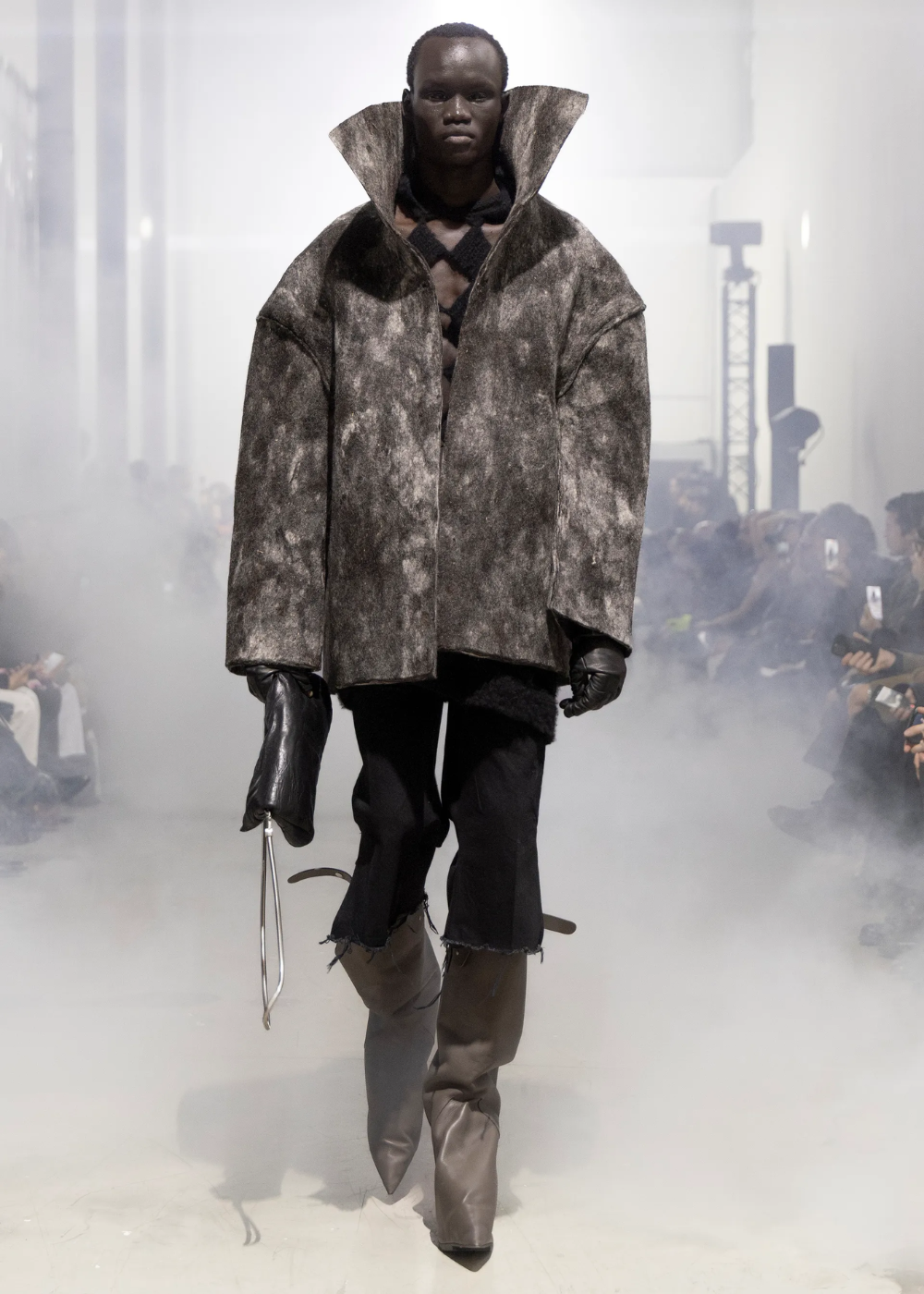 Modelo caminha com postura firme e expressão séria, vestindo casaco oversized cinza texturizado de gola alta e calça preta.