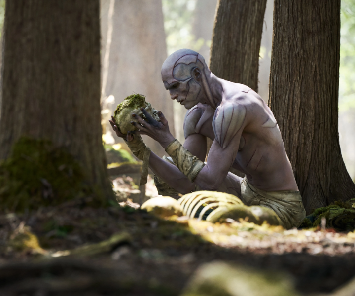 Personagem sem camisa com pintura corporal cinza e detalhes musculares, sentado em floresta, segura crânio coberto de musgo.