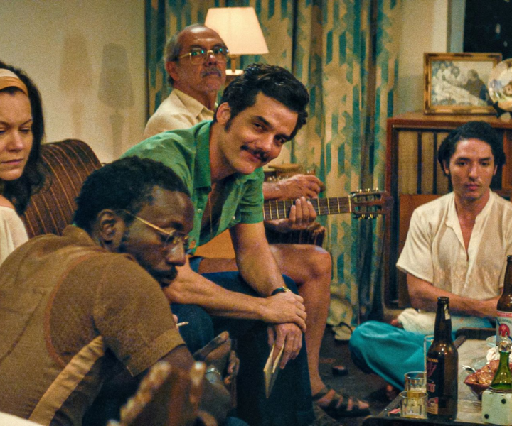 Cena intimista com cinco pessoas em roupas casuais vintage, tons terrosos e verdes dominam ambiente acolhedor.