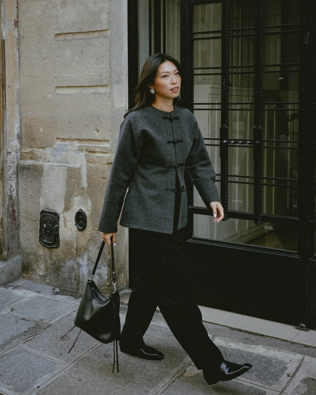 Modelo caminha com casaco cinza de lã com fechamento em nó chinês, calça preta ampla e bolsa de couro, em clima urbano e