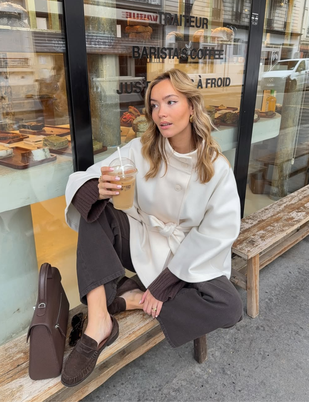 Modelo senta casualmente em banco rústico usando mocassim feminino marrom em camurça, calça ampla preta e casaco branco.