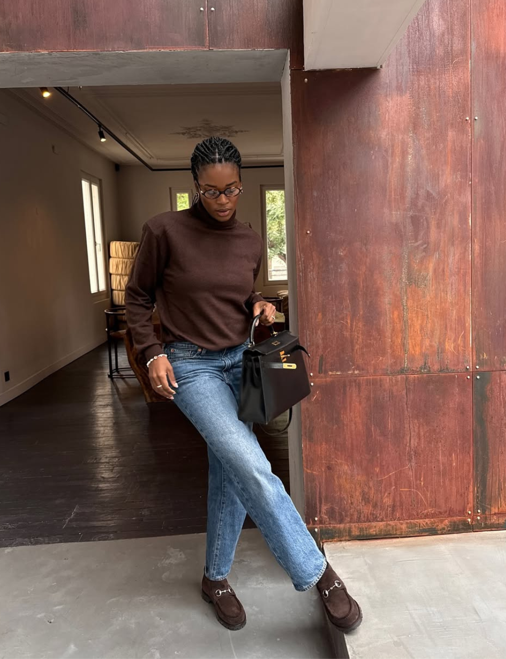 Mulher posa com mocassim feminino marrom em camurça, jeans azul claro e suéter marrom, em ambiente interno aconchegante.