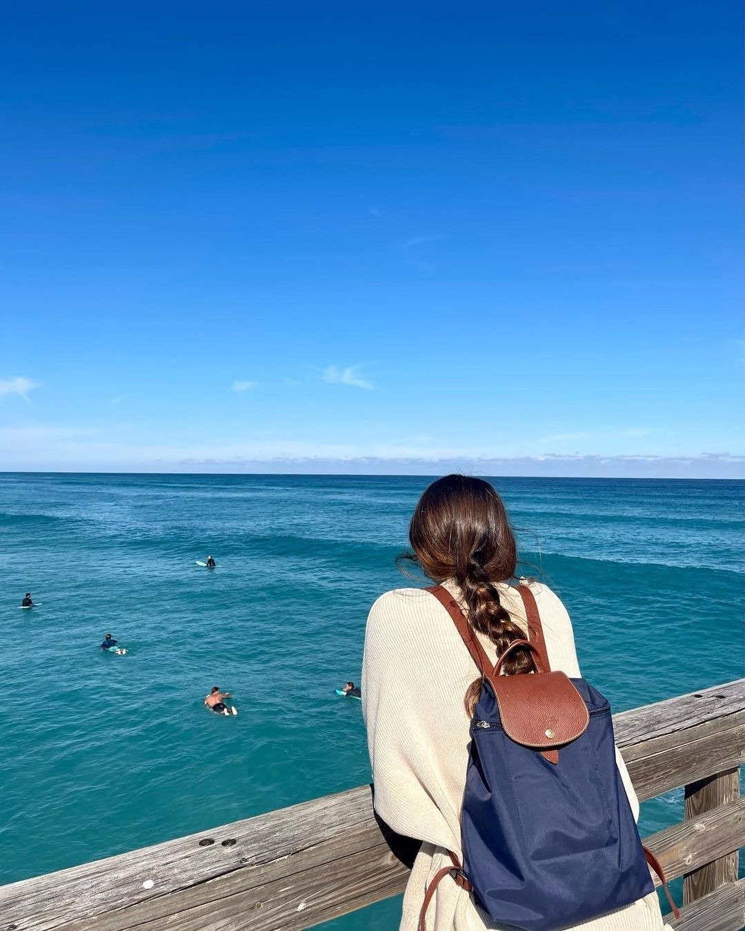 Modelo de costas usa mochila feminina azul marinho com detalhes em marrom e tecido leve, contemplando o mar sob céu claro.