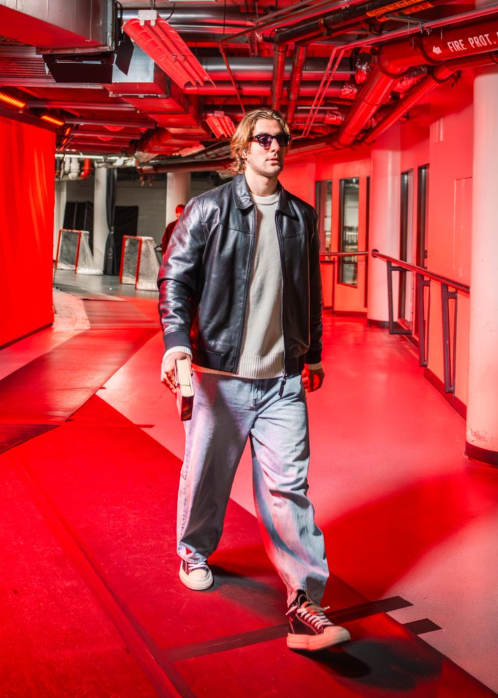 Homem caminha com jaqueta de couro preta, suéter branco e jeans largos, em corredor iluminado por luz vermelha intensa.