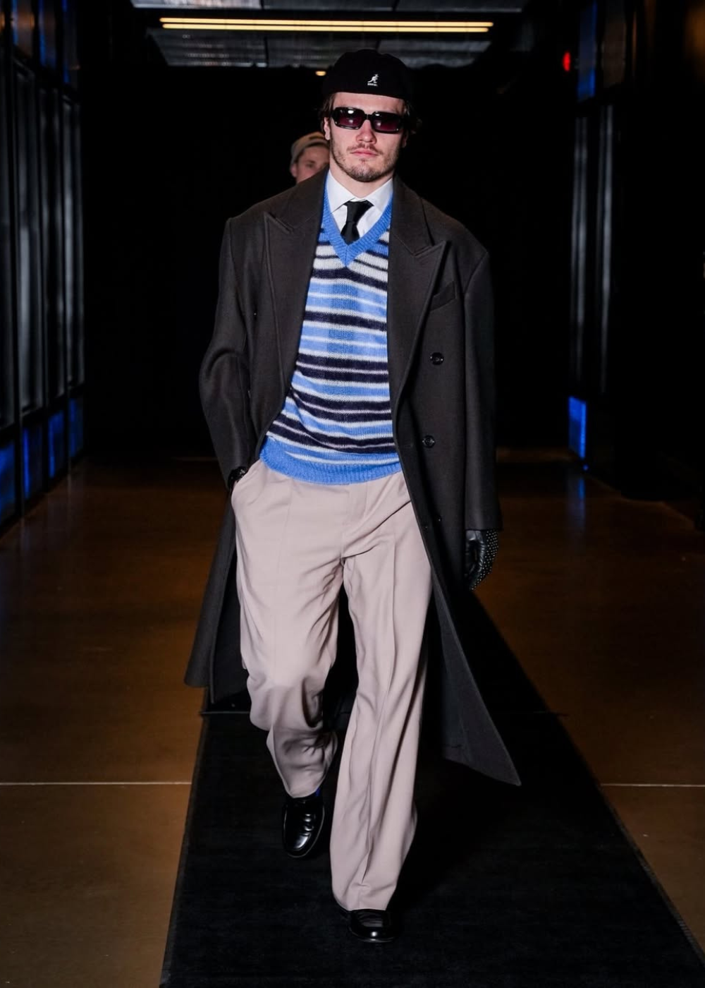 Modelo caminha confiante com look hóquei que mistura suéter listrado azul, calça bege ampla, sobretudo preto e acessórios.