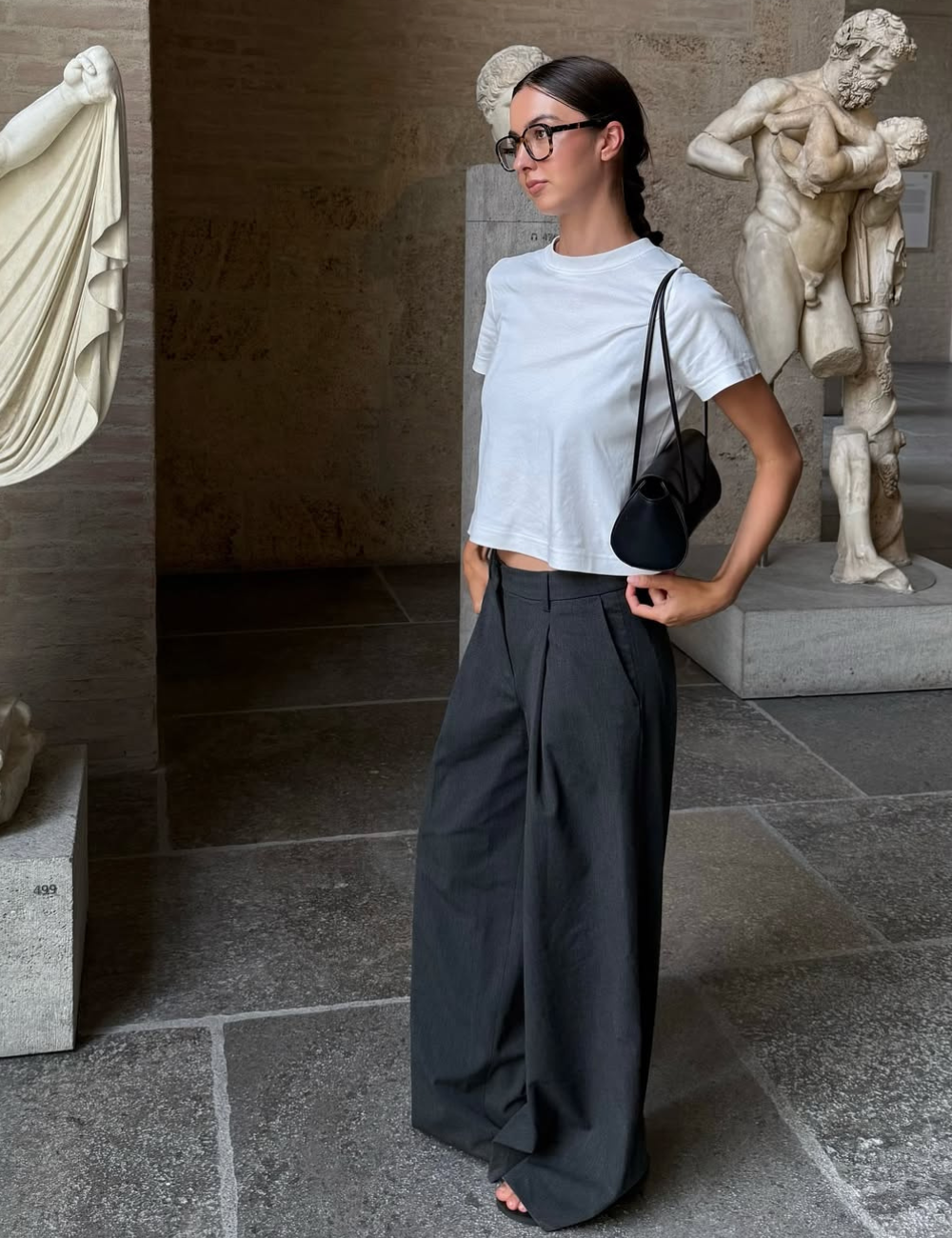 Modelo posa com novos looks minimalistas em camiseta branca cropped de algodão, calça pantalona cinza escura e bolsa preta.