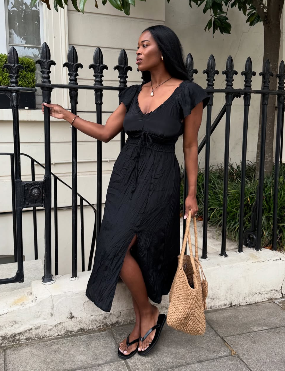 Modelo posa com novos looks em vestido midi preto de tecido leve, mangas bufantes e fenda frontal, combinando bolsa de palha.