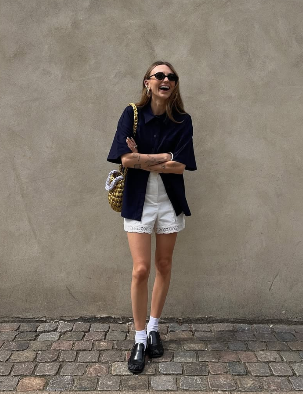 Modelo posa sorridente com camisa azul marinho oversized, shorts brancos de renda, meias brancas e mocassins pretos.