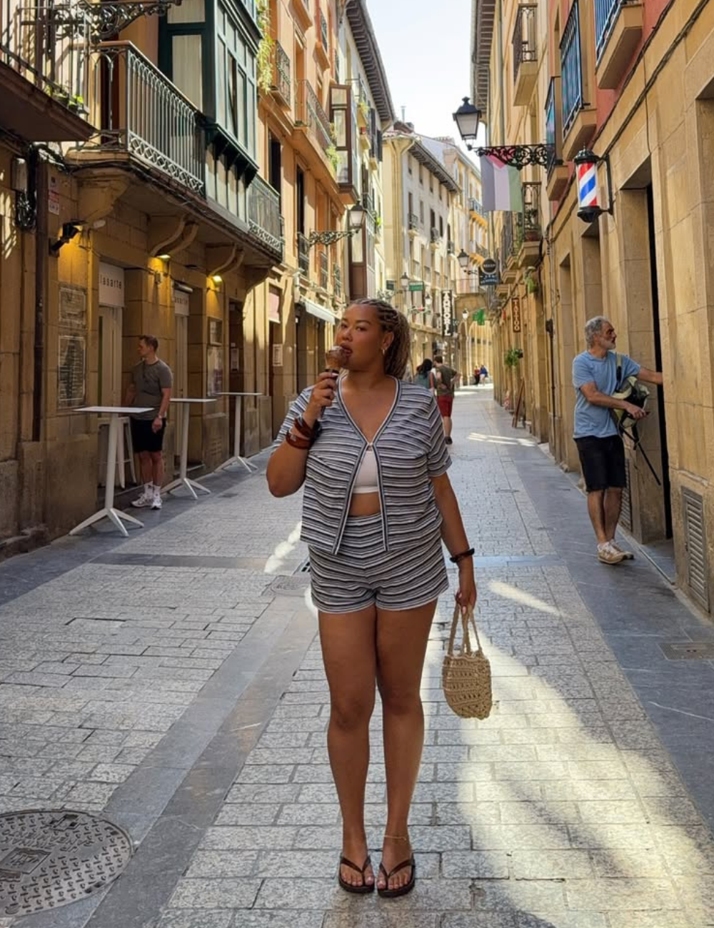 Modelo posa em rua ensolarada com novos looks de shorts e casaco listrados em tons neutros.