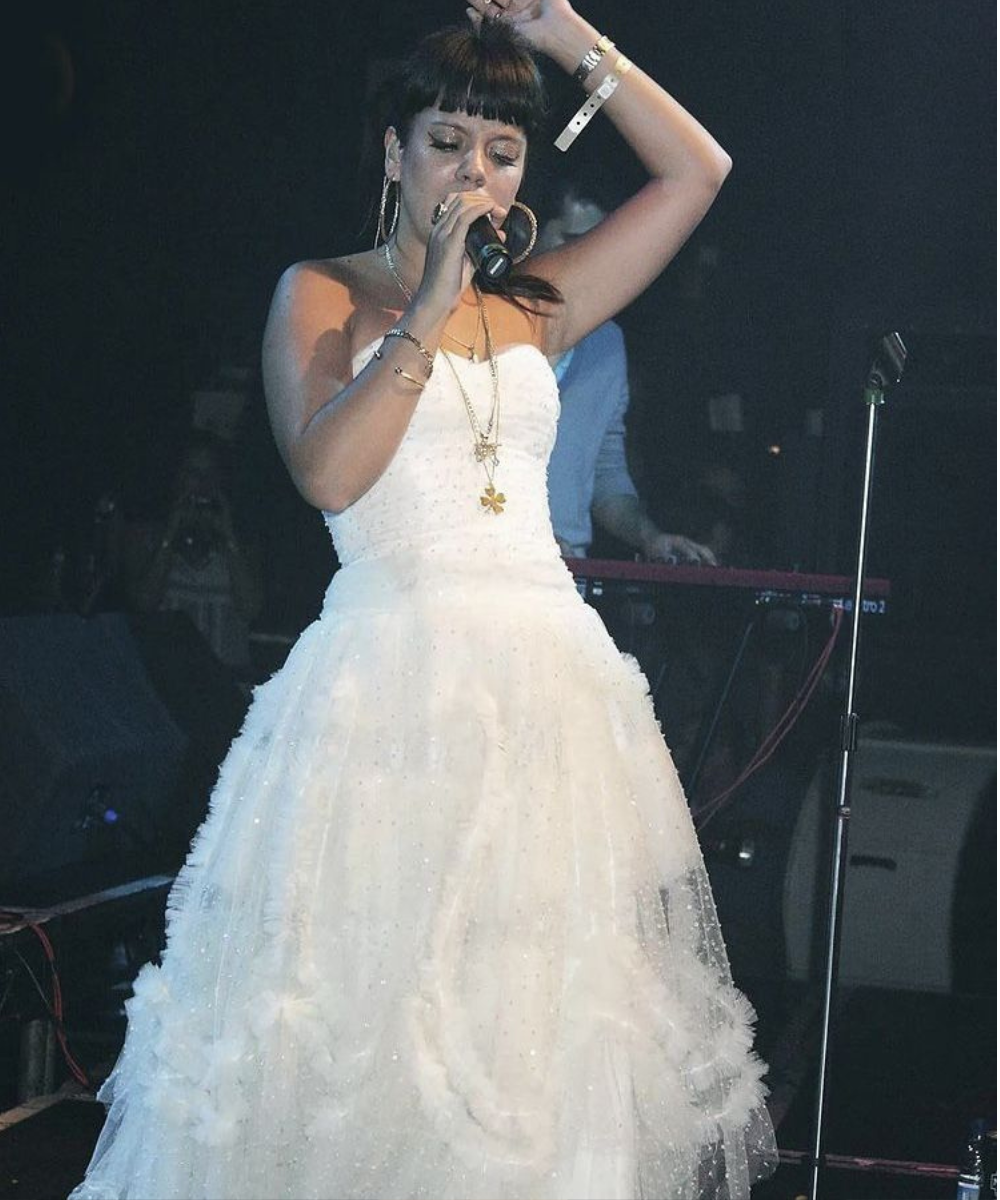 Lily Allen canta com expressão intensa usando vestido branco tomara que caia de tule com texturas delicadas e acessórios