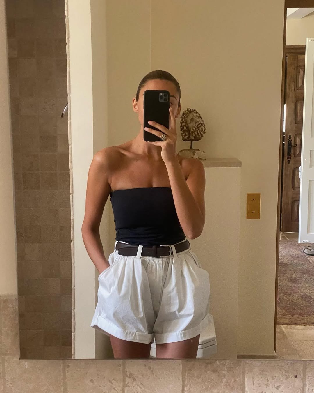 Modelo posa em look minimalista para o calor com top tomara que caia preto e shorts de algodão branco com pregas e cinto