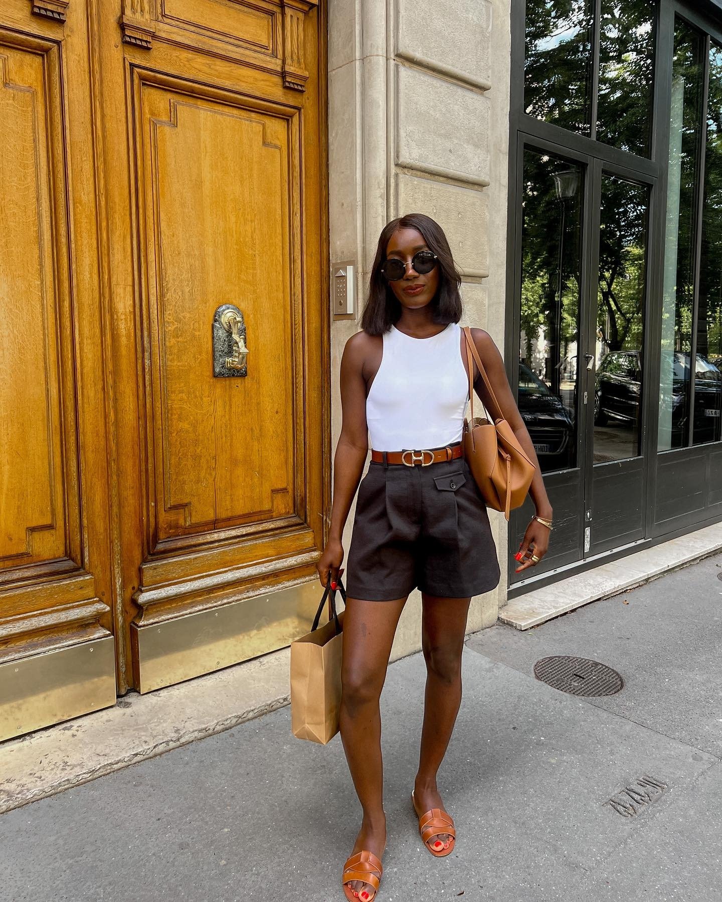 Modelo posa com regata branca justa, shorts pretos de linho com cinto caramelo, sandálias e bolsa em tom terroso, look leve