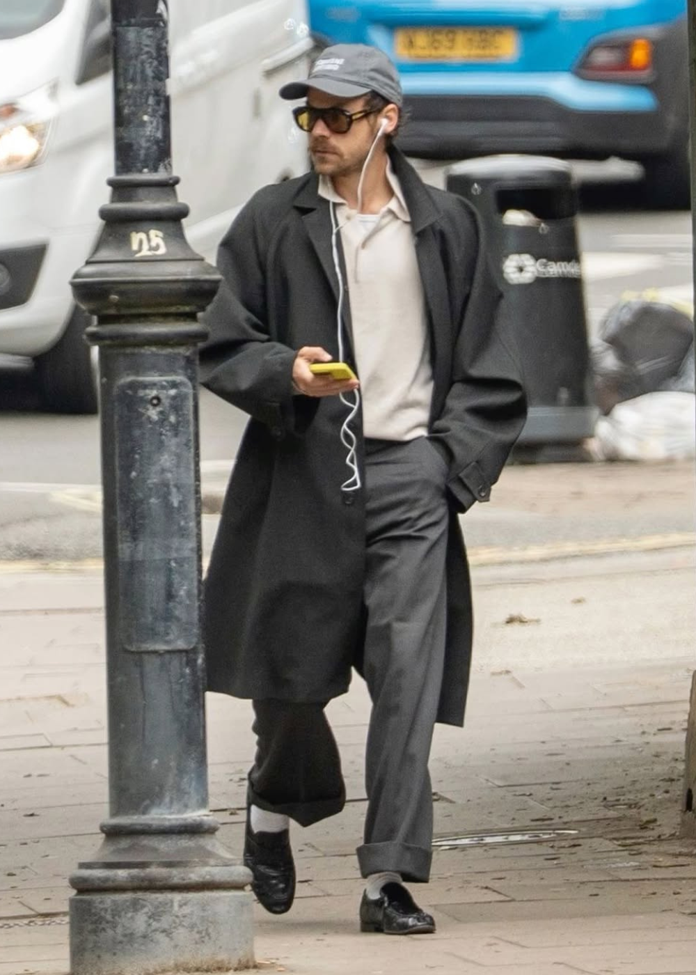 Homem caminha com óculos máscara escuros, sobretudo preto oversized, calça cinza e camisa clara em clima urbano casual.