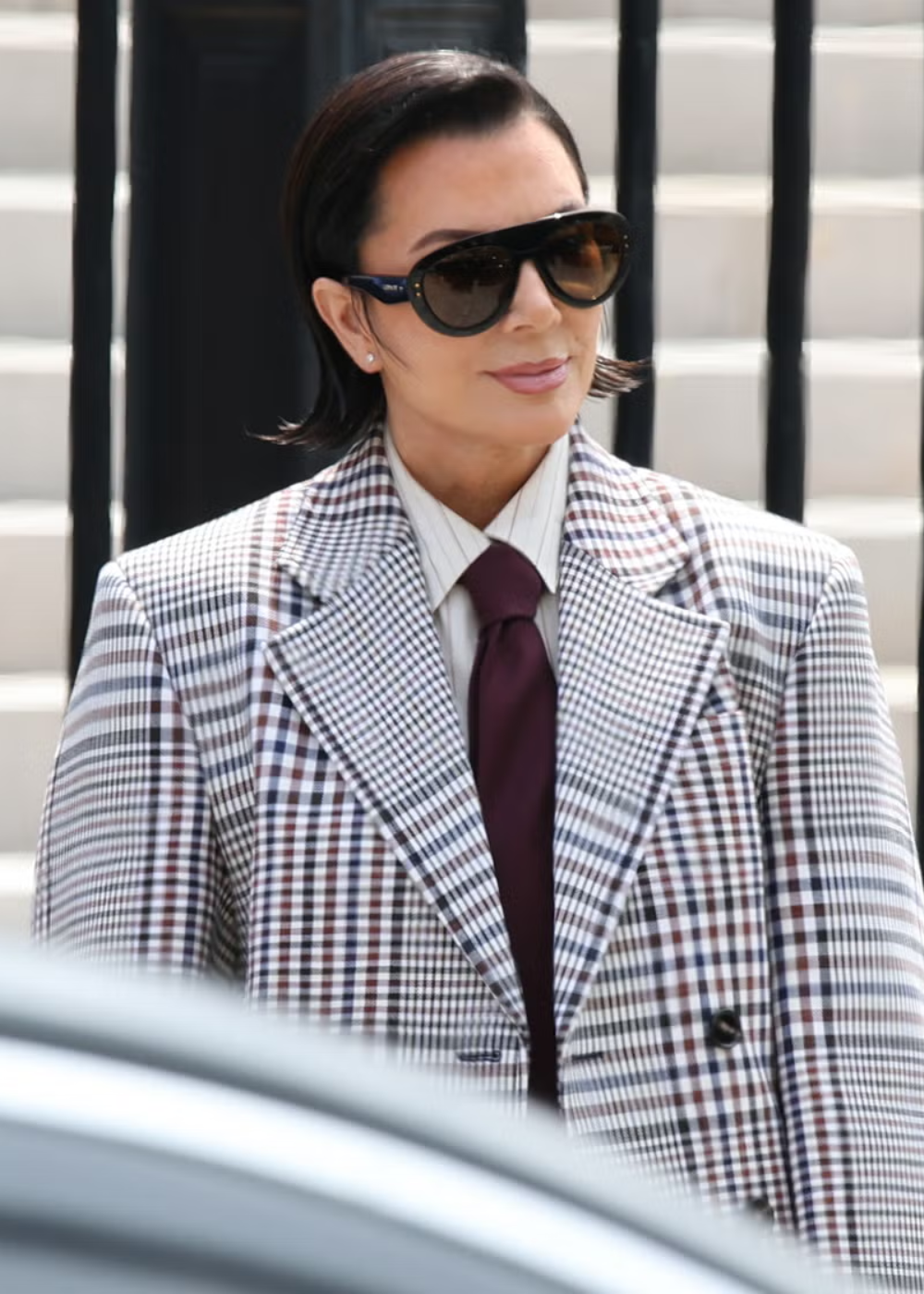 Mulher posa com óculos máscara oversized em acetato preto, blazer xadrez oversized e gravata vinho.