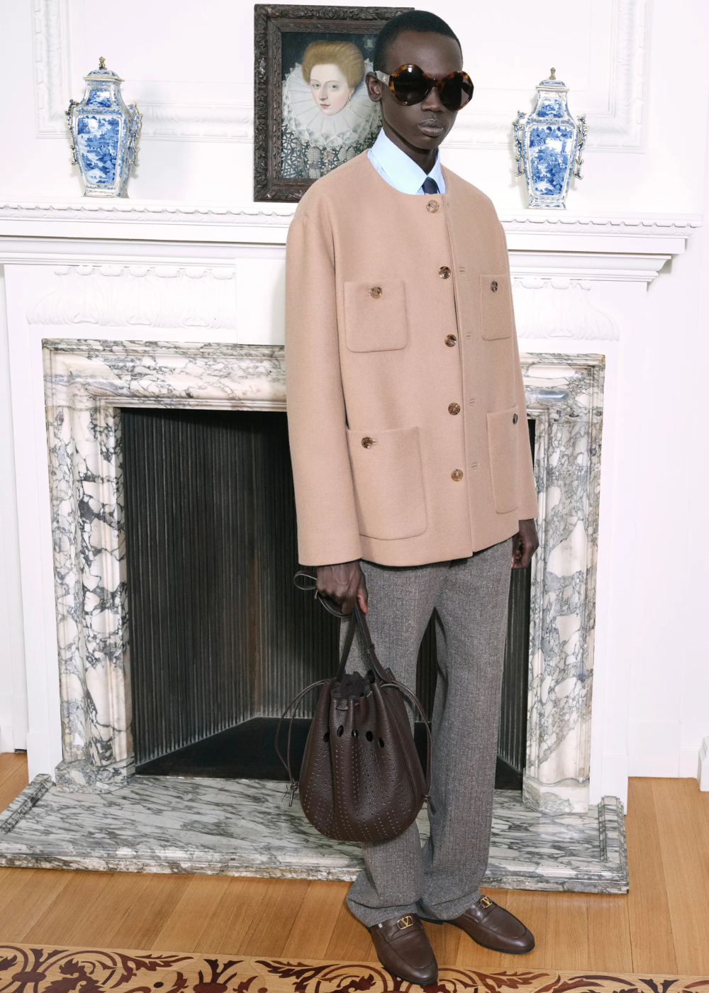 Modelo posa com óculos máscara oversized em acetato tartaruga, casaco bege de lã com bolsos frontais, calça cinza e bolsa.